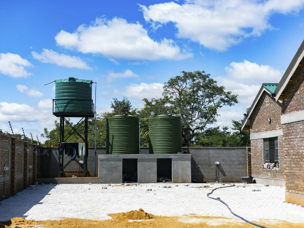 Three green water tanks beside a brick house, one raised on a steel tank stand and two on a concrete base